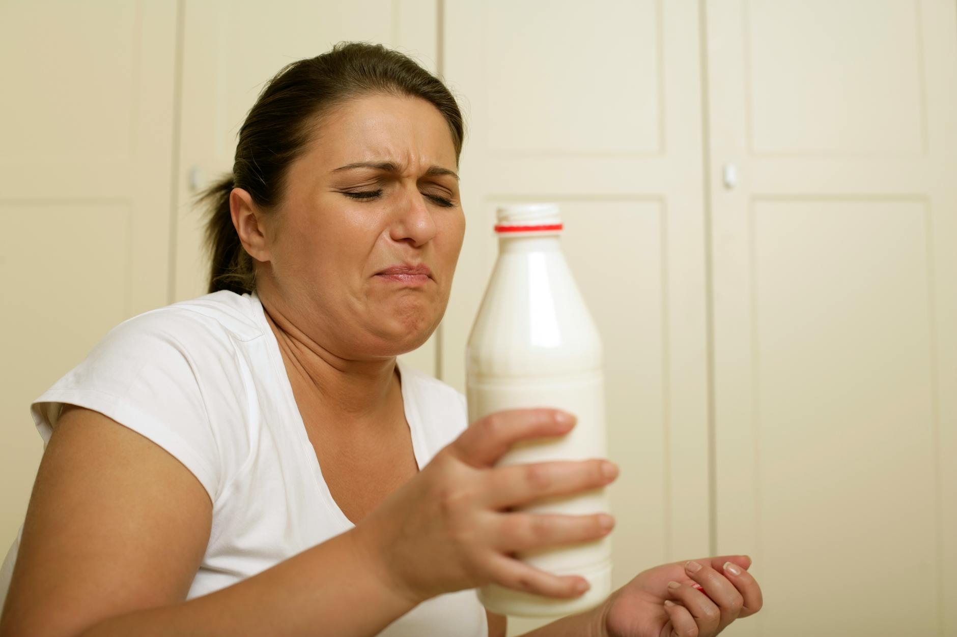 Woman with displeased expression holding a milk bottle indoors, possibly depicting spoiled milk.