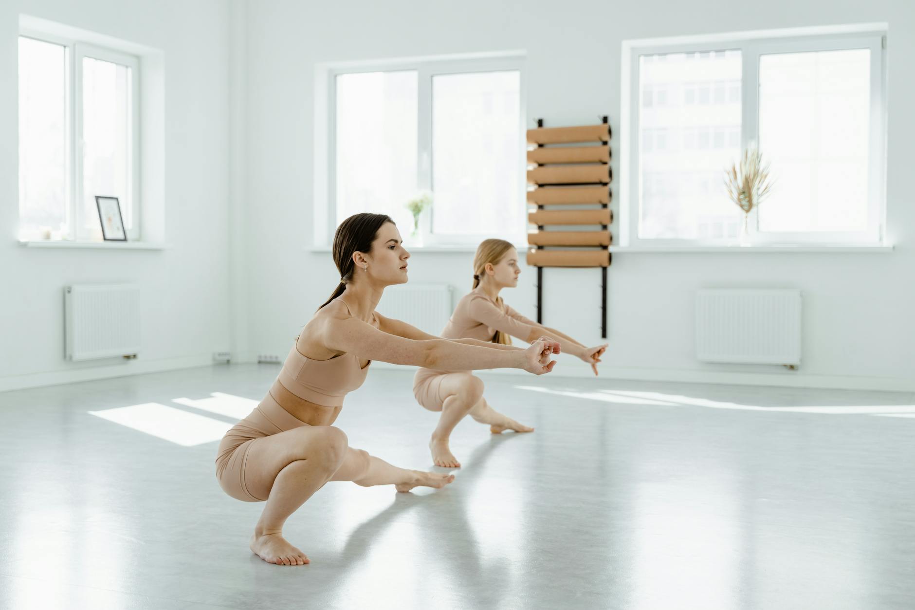Two women performing ballet stretches in an airy, sunlit dance studio.