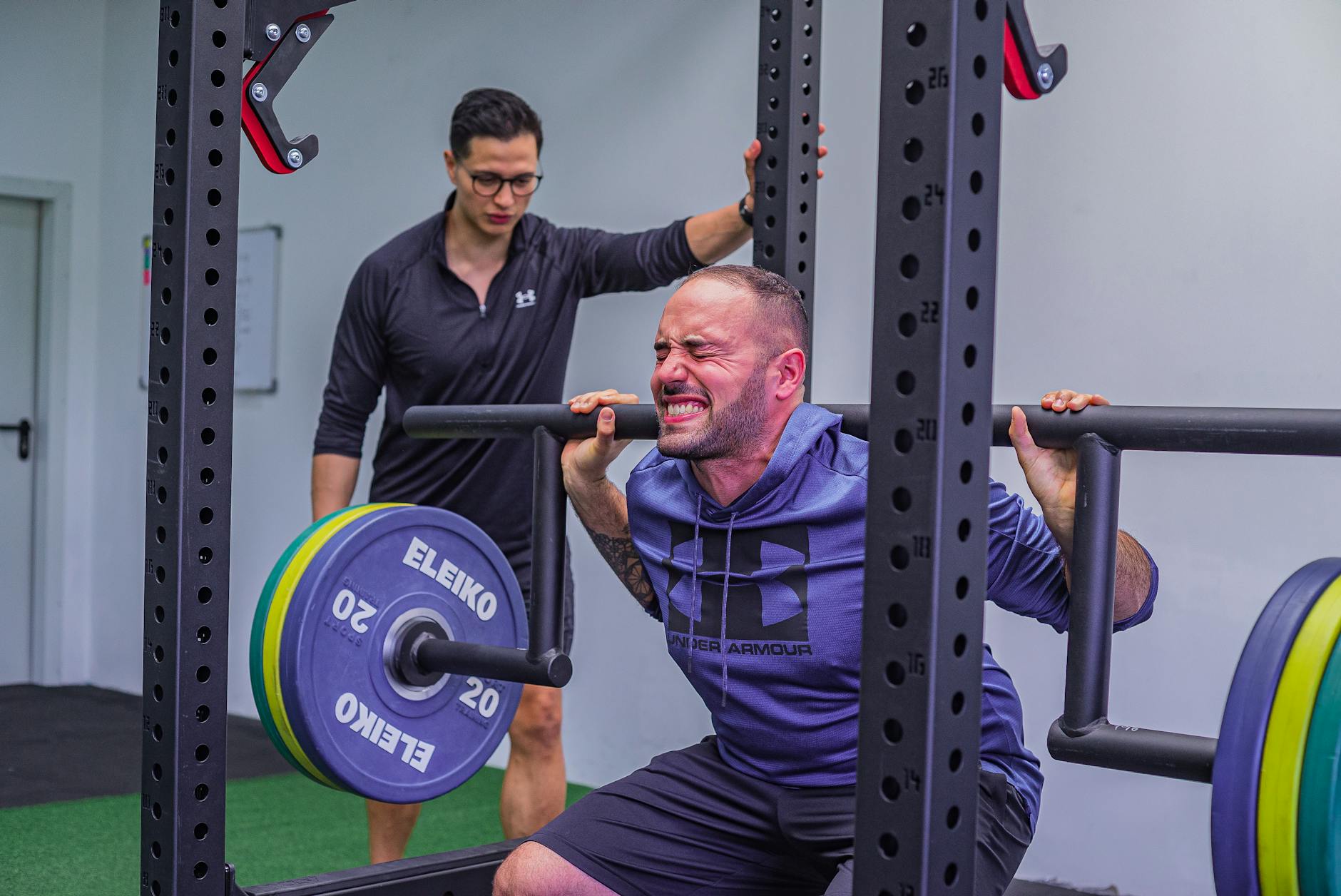 A man intensely performs a squat exercise with a barbell while his coach guides him in the gym.