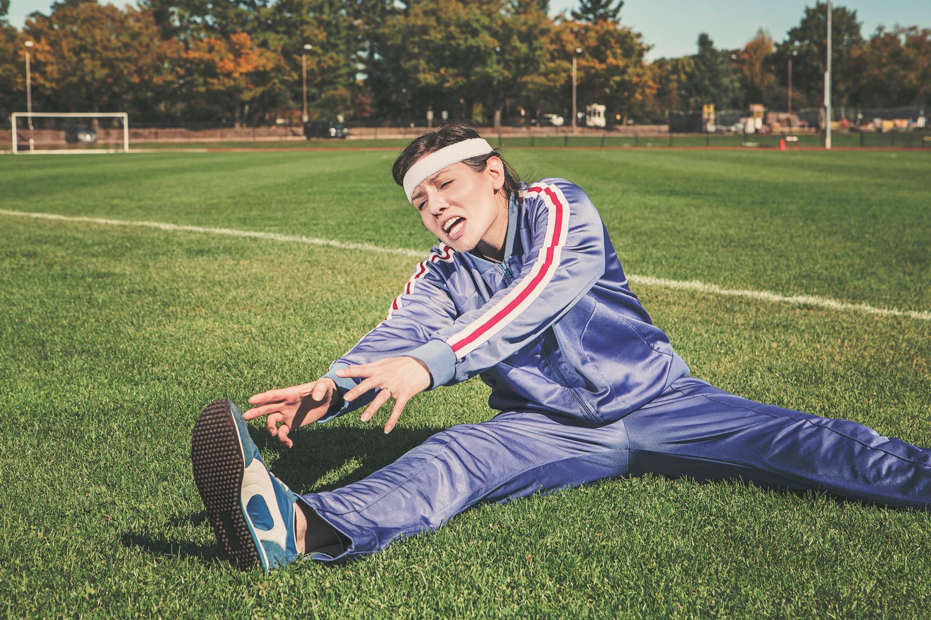 A woman in a tracksuit stretching on a sports field, surrounded by greenery.