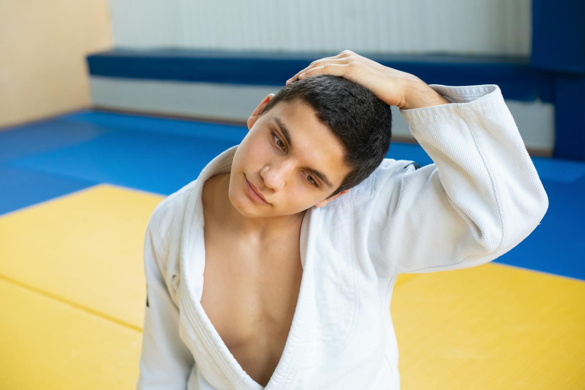 A young man in a judo uniform stretches his neck in a brightly colored gym setting, expressing focus and concentration.